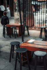 Outdoor seating area with rustic wooden tables and black stools. A young child stands near tables in a blurred background. Urban, minimalist cafe or restaurant setting; vertical layout