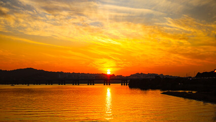 A breathtaking sunset paints the sky in fiery hues of orange and yellow. The sun dips below the horizon, casting a golden glow on the calm river and silhouetting the bridge.