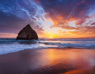 Dramatic coastal sunset with golden light reflecting on wet sand and crashing waves, framed by towering rocky cliff and vibrant orange and blue cloudy sky.