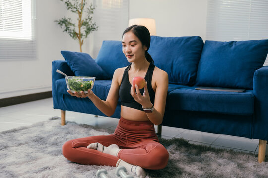 Sitting on a rug in her living room, a young woman in sportswear contemplates her healthy food choices, holding a bowl of fresh salad and an apple, promoting a balanced diet and mindful eating