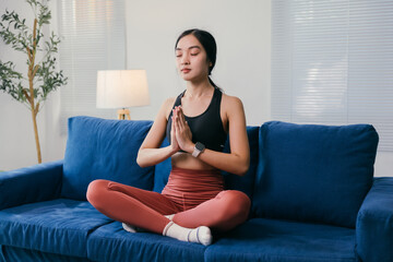 Obraz premium Young asian woman wearing sportswear is practicing yoga and meditation on a blue sofa in her living room, promoting a healthy and mindful lifestyle at home