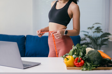 Young woman measuring her waist with a tape measure while standing near a table with fresh vegetables and a laptop, promoting a healthy lifestyle at home