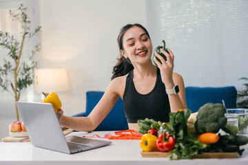 Young smiling asian nutritionist holding bell peppers and following a personalized online diet plan on a laptop, promoting healthy eating habits and weight management through technology
