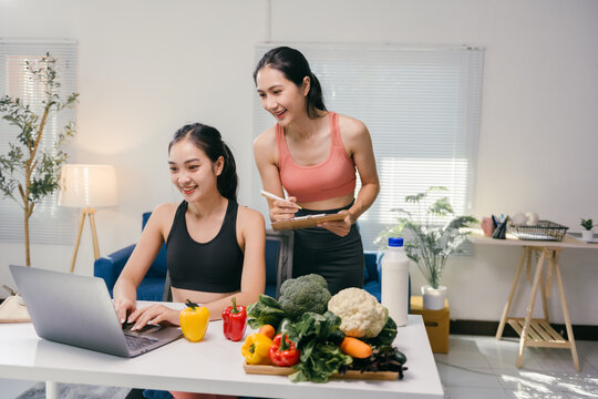 Two smiling women creating a personalized diet plan using a laptop, with fresh vegetables and milk on the table, promoting healthy eating habits and online nutrition consultations