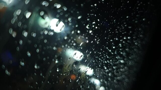 Rain at night on the side of the highway seen from the perspective of inside the car, you can see raindrops sticking to the car window. There is light from street lights, cars and other vehicles passi