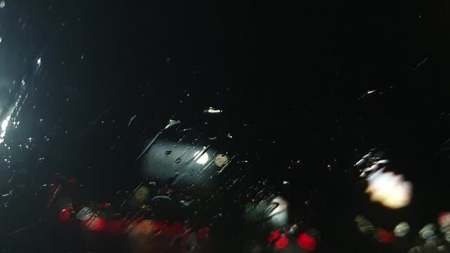 Rain at night on the side of the highway seen from the perspective of inside the car, you can see raindrops sticking to the car window. There is light from street lights, cars and other vehicles passi