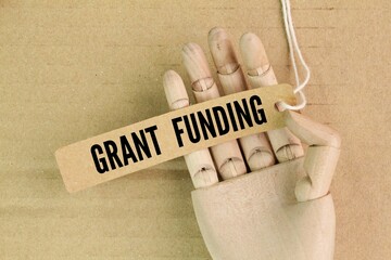 wooden hand and paper tag with the word GRANT FUNDING. any funding or other aid or assistance from...