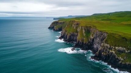 Aerial view of green cliffs meeting a calm blue sea under a cloudy sky, showcasing natural coastal beauty.