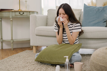 Sick Asian woman sitting on living room and sneezing into tissue paper, ill young female cough and blowing her nose. Health care and medicine concept.