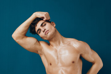 Confident young man with curly hair posing shirtless against a teal background, showcasing a strong...