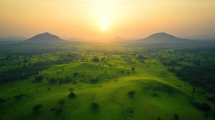 Serene tropical landscape at sunrise, with mist gently hovering over lush green fields and scattered trees, framed by distant mountains.