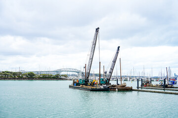 Industrial cranes working on a floating platform in a calm harbor with boats and sailboats in the background during an overcast day
