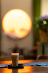 a glass of iced coffee on a tray with a straw, placed on a table with warm lighting. A large, glowing circular light in the background creates a cozy, sunset-like ambiance.