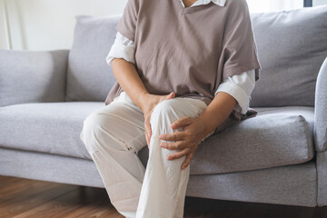 Body arthritis muscles stiff problem, aged woman suffering on leg, pain knee from disease chronic, hand massage rubbing joint injury, mature hurt or sore sitting on sofa at home. Health care people.