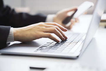 Picture focuses on a woman's hands typing on a contemporary laptop keyboard, office setting appearing blurred