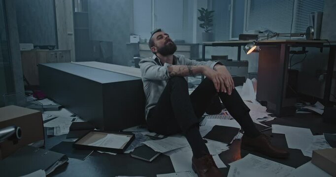 Dolly shot of depressed businessman sitting on floor after fit of rage over failed business deal or receiving bad news. Stressed office employee in destroyed office room. Financial crisis or firing.