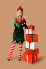 Young woman in elf costume with Christmas gift boxes blowing kiss on brown background