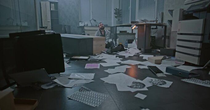 Dolly shot of depressed office worker sitting on the floor in destroyed office room. Frustrated businessman after fit of rage over unsuccessful business deal or receiving bad news. Burnout at work.