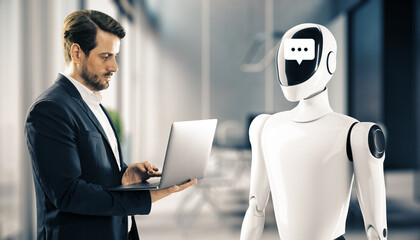 Businessman looking at laptop while standing in front of a humanoid robot with a chat symbol on its...