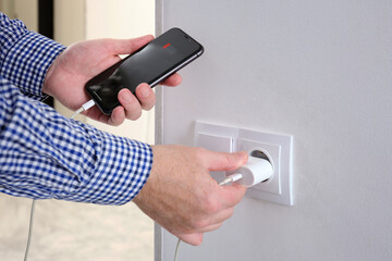 A man holds a smartphone with low battery and plugs the charger into a socket in a living room.
