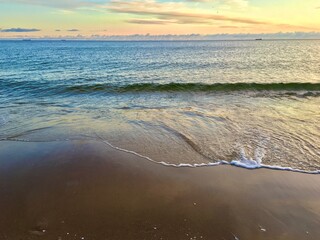Calm sea waves gently washing over the sandy shore during sunset, with a colorful horizon blending soft blues, oranges, and yellows, creating a tranquil coastal scene.