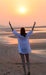 woman walking toward the sun along the shoreline by the sea wearing a white shirt in the morning light