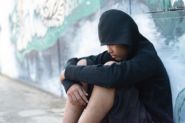 Asian male teenager wearing black headscarves sits on the outside wall of a building as he was punished by his parents for bad behavior, which is a problem in today's society, homeless people concept.