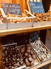 Various types of mushrooms in crates and displayed at a farmer’s market.  Chalkboard signs showing the names of the mushrooms and prices in British Pounds. 