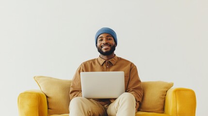 A man wearing a blue hat is sitting on a couch with a laptop in front of him