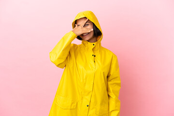 Young latin woman wearing a rainproof coat over isolated background covering eyes by hands and smiling