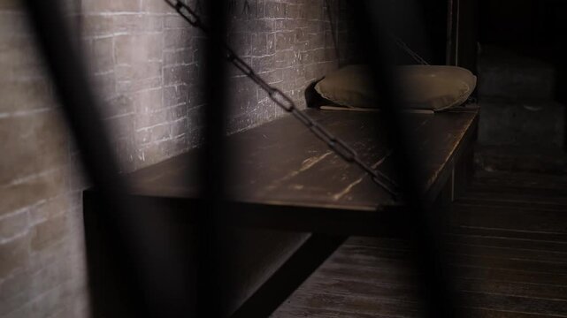 A close-up of a wooden bed hanging on chains, a shelf with a pillow in an old prison cell. A dark room with a solid wooden bed hanging on chains. An old prison.