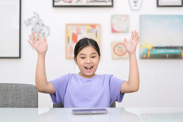 asia child happy excited on smart phone or kid girl fun playing mobile phone or young people looking smartphone on white table and raise hands to wow open mouth with art picture frame background