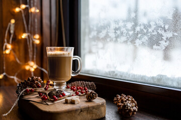 Christmas composition with hot cocoa, fir branches and decorations on the windowsill, snow and frosty patterns outside the window