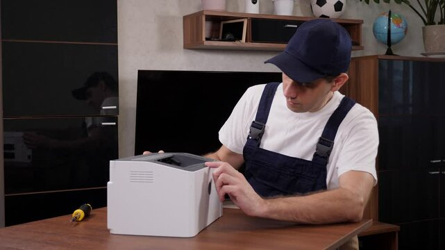 A male handyman in blue overalls inspects a printer before starting repairs in a client's apartment.