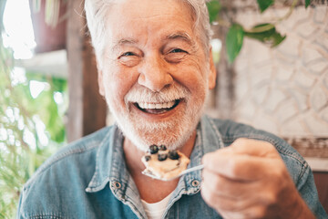 Close up portrait of happy bearded senior man eating a fruit cake in coffee shop enjoying break or...