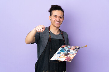 Young artist man holding a palette over isolated purple background points finger at you with a...