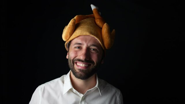 A young Caucasian man in a white shirt smiles at the camera while wearing a turkey hat, celebrating Thanksgiving