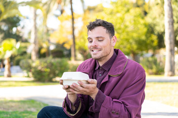 Young caucasian man at outdoors taking a box of takeaway food with happy expression