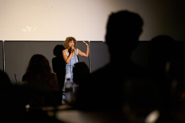 Caucasian female performer with curly hair delivering a monologue on stage in a dimly lit theater...