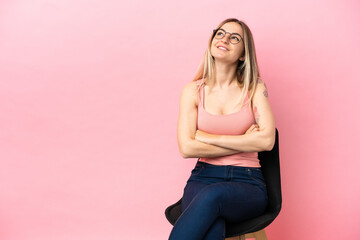 Young woman sitting on a chair over isolated pink background looking up while smiling