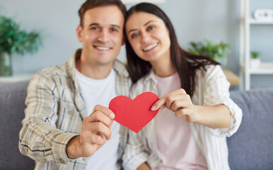 Young cheerful smiling brunette woman and man holding paper heart in hands sitting on sofa at home and looking at camera. Happy couple in love. Romantic, relationship and Valentines day concept.