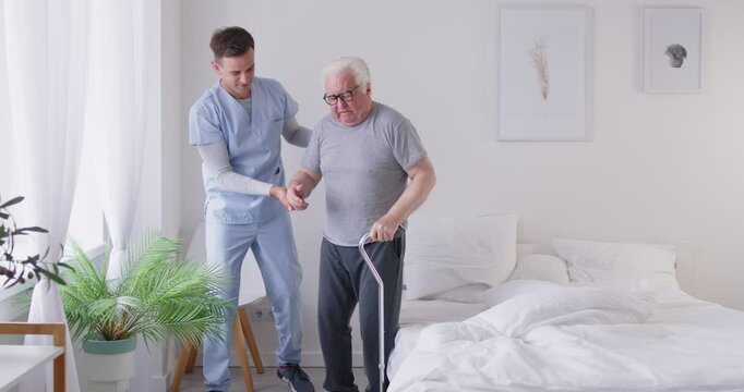 Doctor or nurse helps a senior male patient walk with a stick during a rehabilitation session in a nursing home. The healthcare professional provides support and care during the patient recovery.