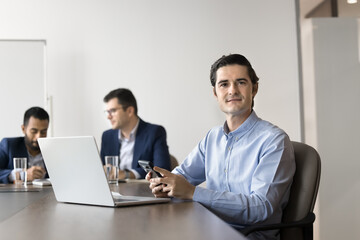 Positive confident business leader man using smartphone and laptop, sitting at work meeting table with team of colleagues behind, looking at camera for professional portrait