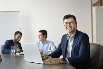 Happy handsome middle aged Caucasian business leader man in glasses, using laptop in meeting room, sharing large table with diverse team, looking at camera for professional portrait, smiling
