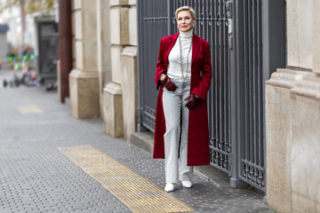 Stylish Mature Woman in Trendy Burgundy Coat Standing City, Wearing Fashionable clothing White Turtleneck Sweater, Pearl Necklace and Grey Jeans. Confident and Chic Fashion Model Street Style