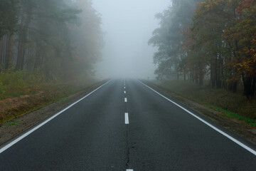 Asfaltowa droga prowadząca przez las, w mglisty, jesienny poranek. Nad jezdnią unosi się mgła ograniczająca mocno widoczność. © boguslavus