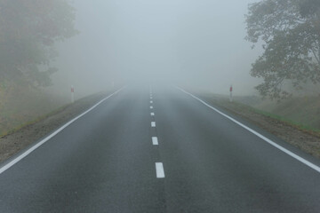 Asfaltowa droga prowadząca przez las, w mglisty, jesienny poranek. Nad jezdnią unosi się mgła ograniczająca mocno widoczność. © boguslavus