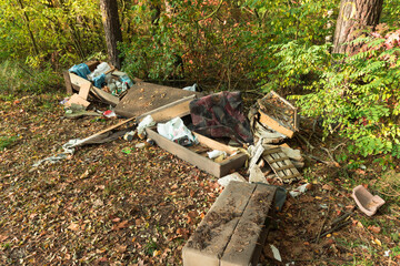 Porzucone w lesie odpady. Na tle drzew widać leżące na warstwie opadłych, brązowych liści elementy kanapy, puszki, butelki plastikowe będące odpadami.
