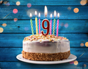 Ninth birthday cake with white frosting and nut crust, decorated with colorful striped candles and number 9, set against a blue wooden backdrop with festive bokeh lights