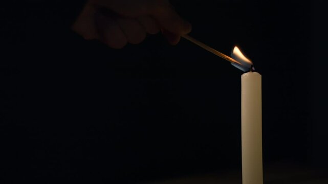 Male hand lighting a white thin taper candle using a safety match on a black background, close up shot. Grief, loss, and melancholy concepts.